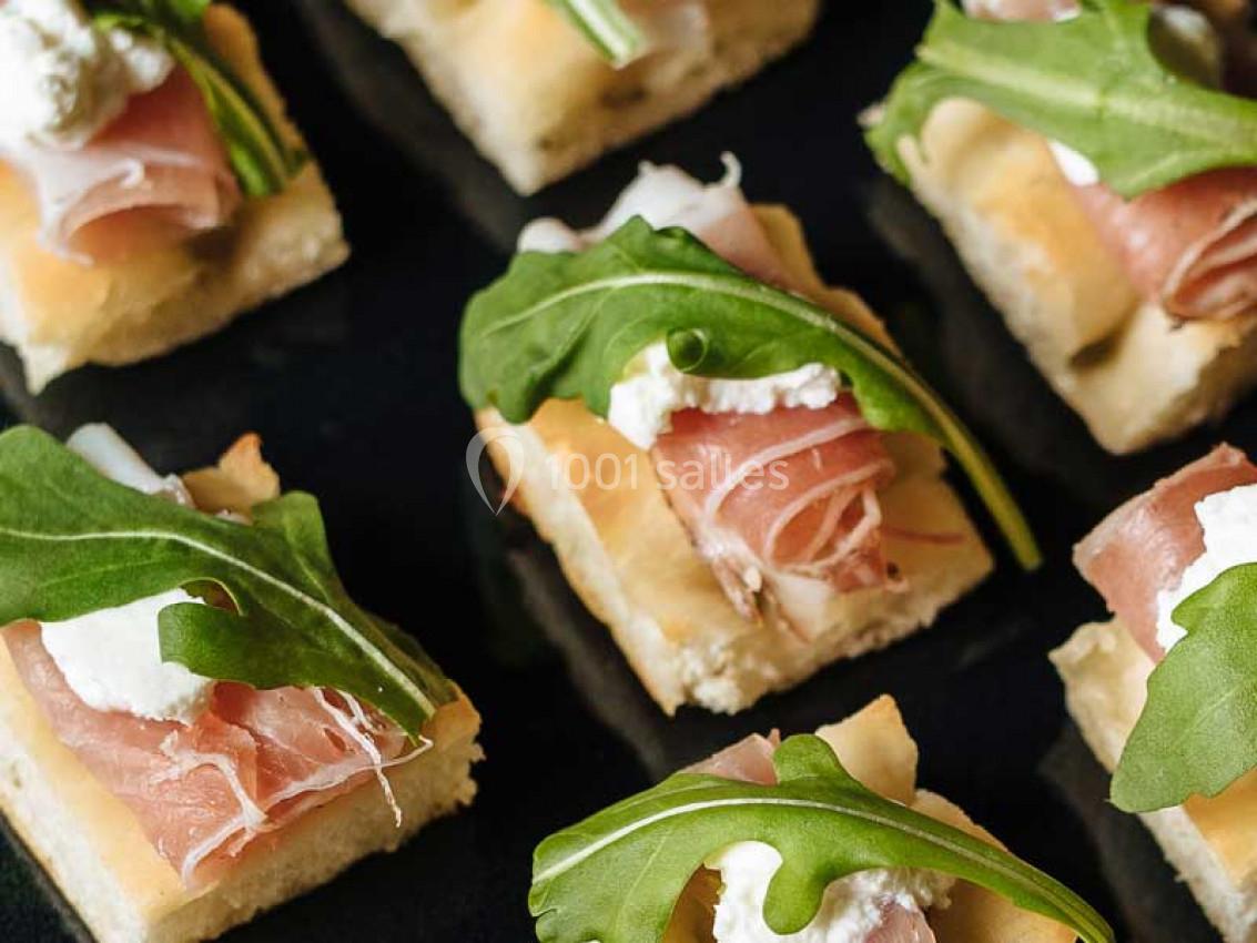 Carrés de focaccia garnis de jambon cru, fromage frais et feuilles de roquette sur une surface noire.