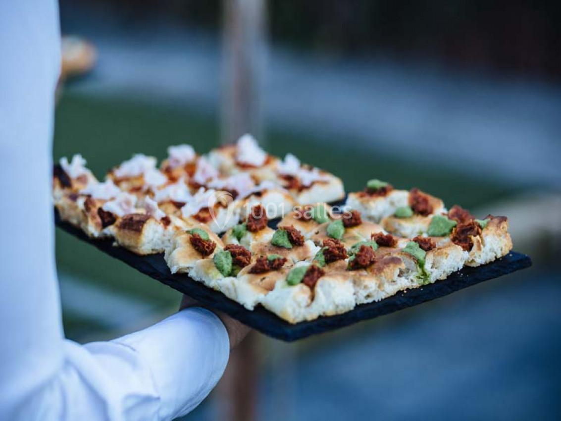 Plateau de focaccia garnie de morceaux de viande, herbes fraîches et fromage, tenu par une personne en chemise blanche.