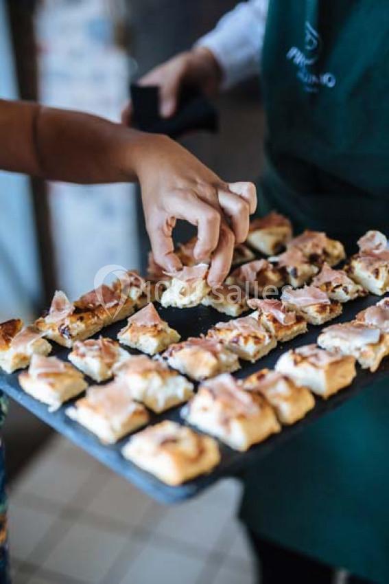Une main prend un morceau de focaccia garnie de jambon sur un plateau tenu par une personne en tablier vert.