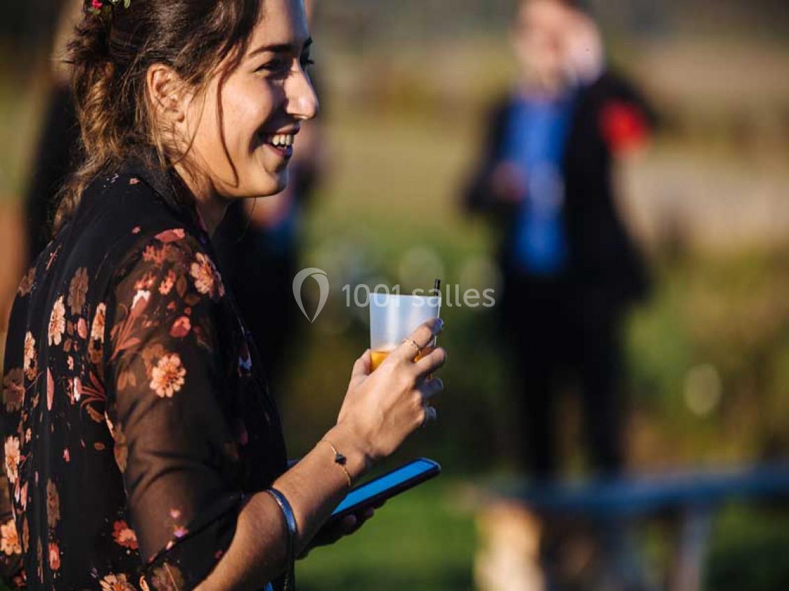 Femme souriante tenant un verre et un téléphone dans un espace extérieur ensoleillé avec des personnes en arrière-plan flou.