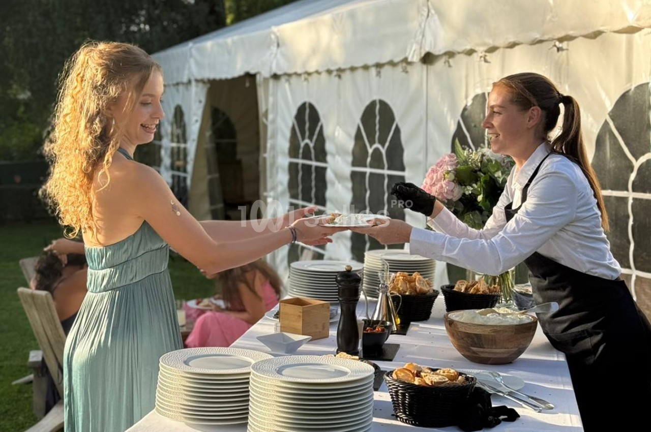 Une femme en robe reçoit une assiette d'une serveuse devant un buffet en plein air près d'une tente blanche.