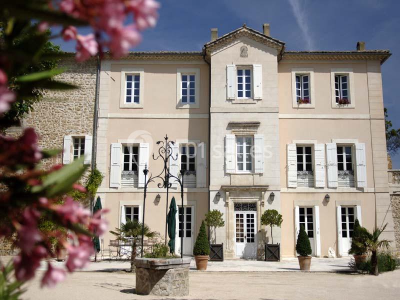 Façade d'une maison en pierre à deux étages avec volets blancs, entourée de plantes et d'une cour ensoleillée.