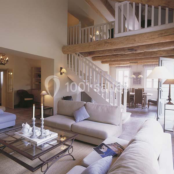 Salon lumineux avec poutres apparentes, canapé beige, table basse en verre et escalier menant à une mezzanine.