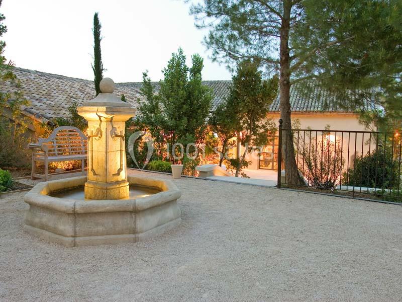 Fontaine en pierre éclairée dans une cour gravillonnée entourée de végétation et d'une maison en arrière-plan.