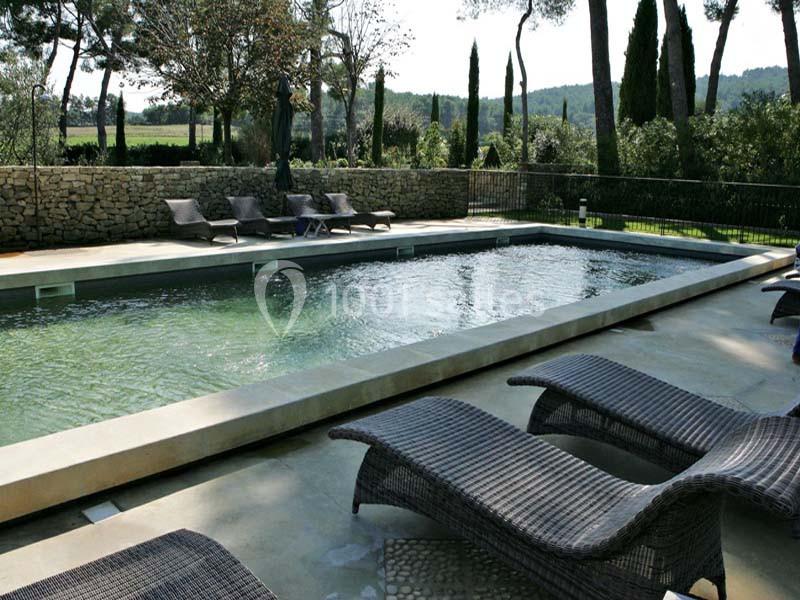 Piscine rectangulaire extérieure entourée de chaises longues en rotin, avec vue sur des arbres et un mur en pierre.