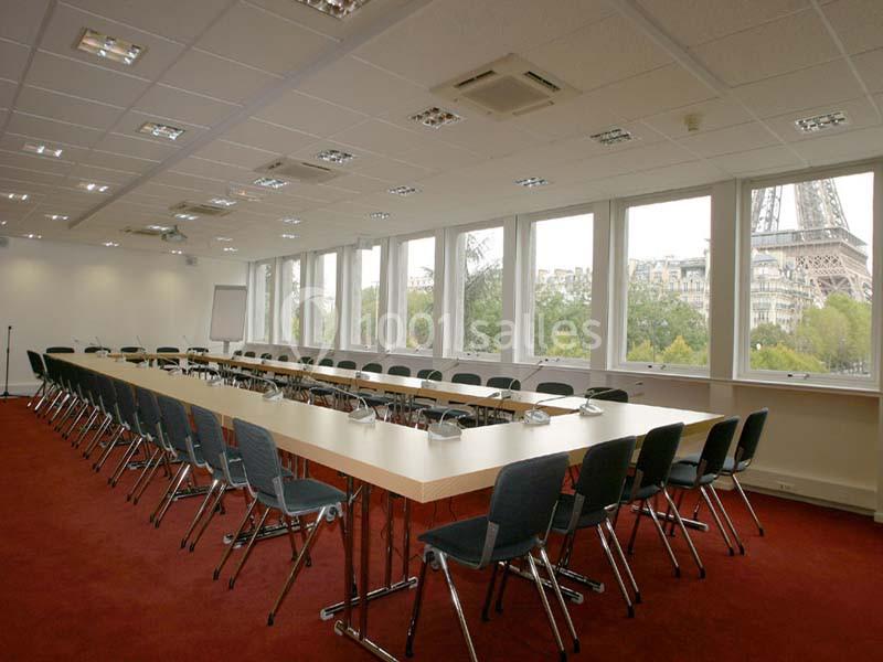 Salle de réunion spacieuse avec de longues tables en U, chaises alignées et vue sur la Tour Eiffel par les fenêtres.