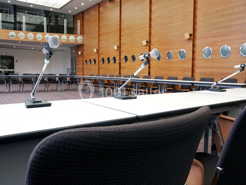 Salle de conférence avec des micros sur des tables blanches, entourée de murs en bois et de sièges noirs.