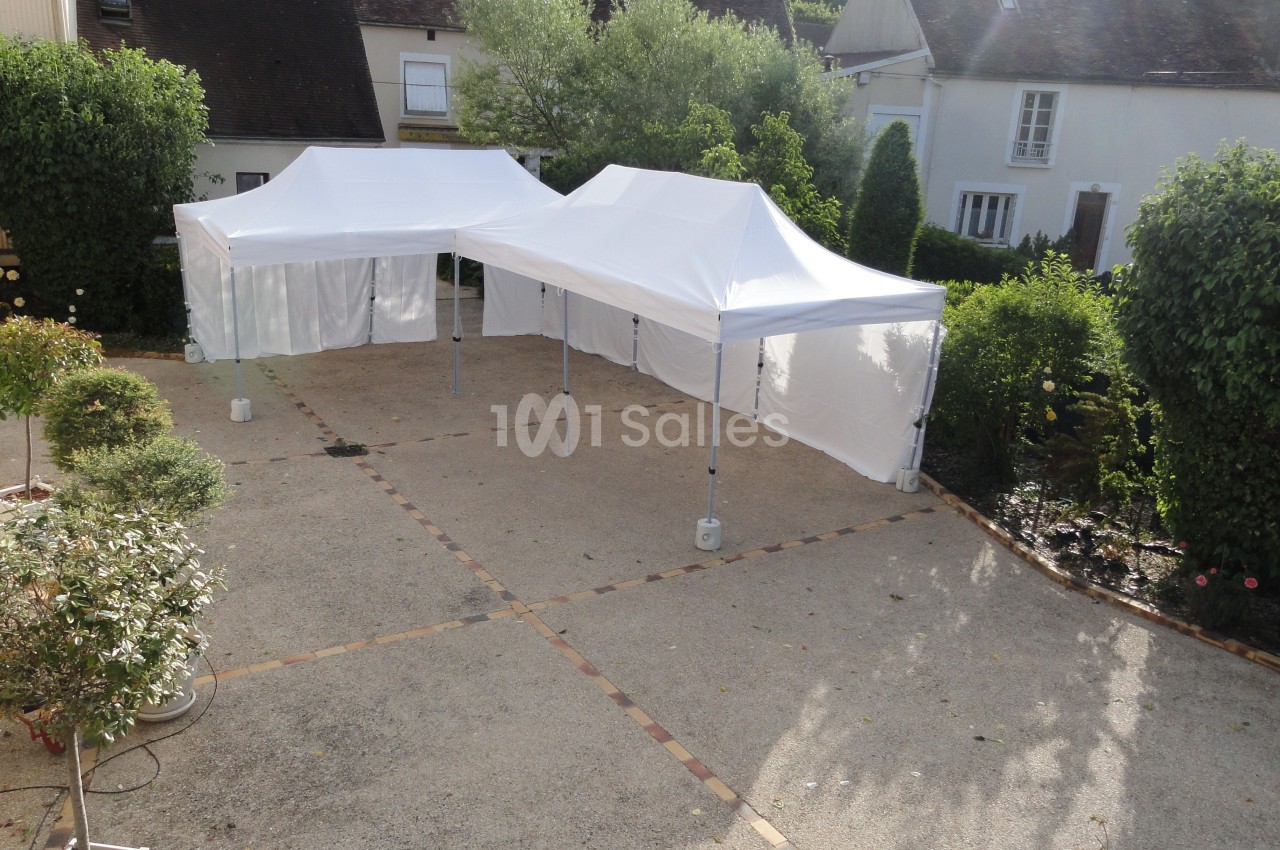 Deux tentes blanches montées sur une terrasse pavée entourée de végétation et de maisons.