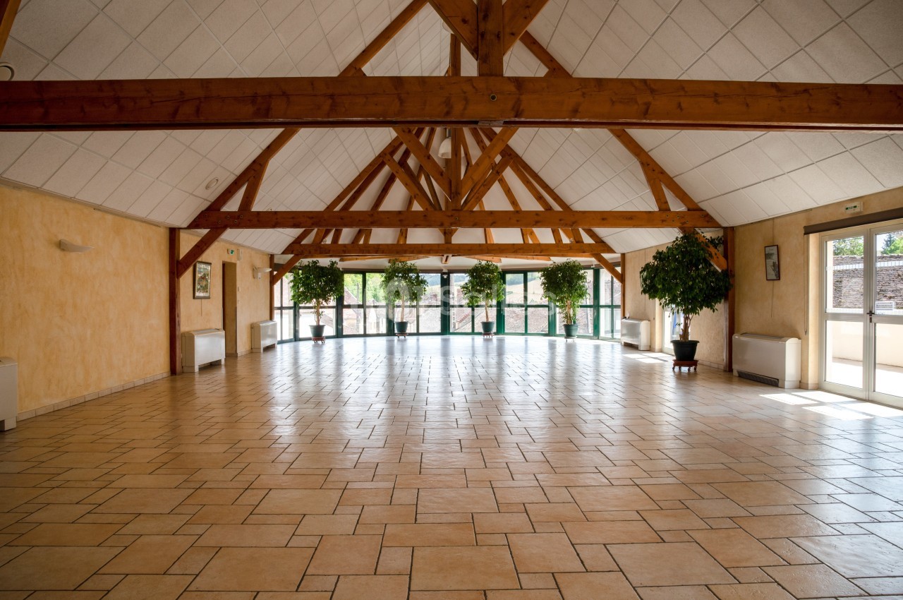 Grande salle lumineuse avec charpente en bois apparente, carrelage clair et grandes baies vitrées donnant sur l'extérieur.