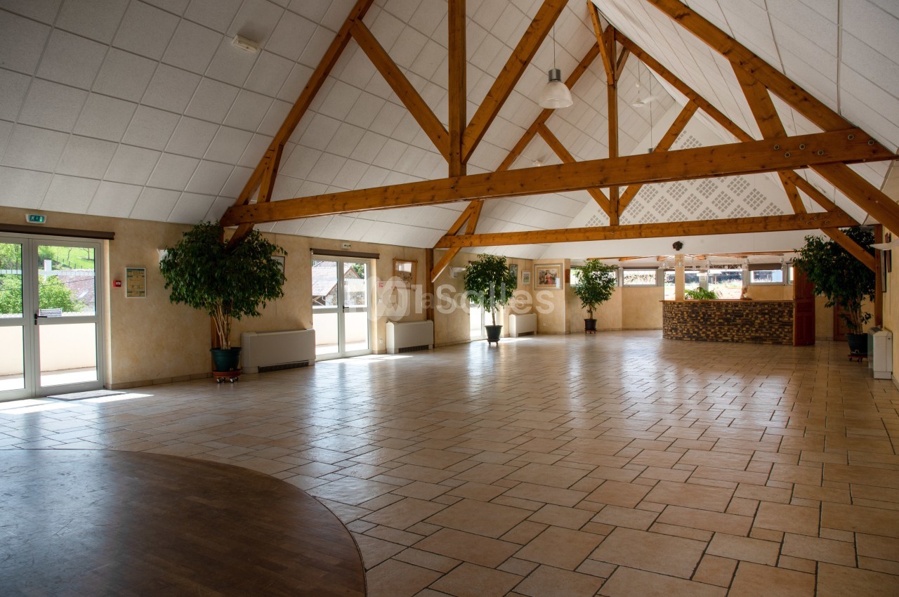Salle spacieuse avec charpente en bois apparente, sol carrelé, grandes fenêtres et comptoir en pierre au fond.