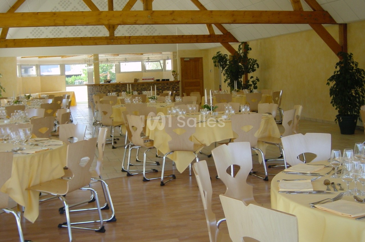 Salle de réception lumineuse avec tables rondes dressées, nappes jaunes et chaises en bois clair sous une charpente…
