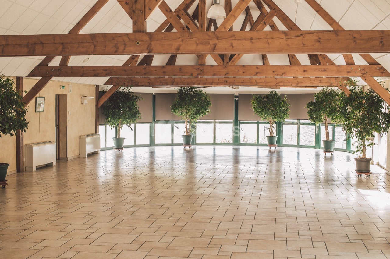 Salle spacieuse avec charpente en bois apparente, carrelage clair et plantes en pots alignées près des fenêtres.