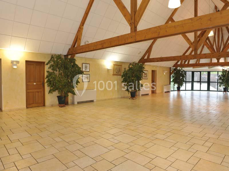 Grande salle lumineuse avec carrelage clair, poutres apparentes en bois, murs beige et plantes en pot.