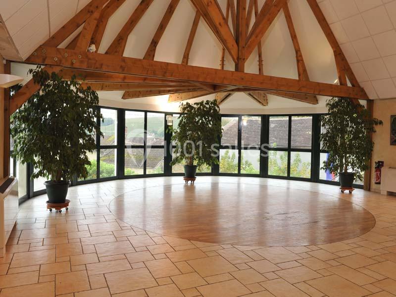 Salle lumineuse avec charpente en bois apparente, grandes baies vitrées et sol en carrelage beige, décorée de plantes en pot.