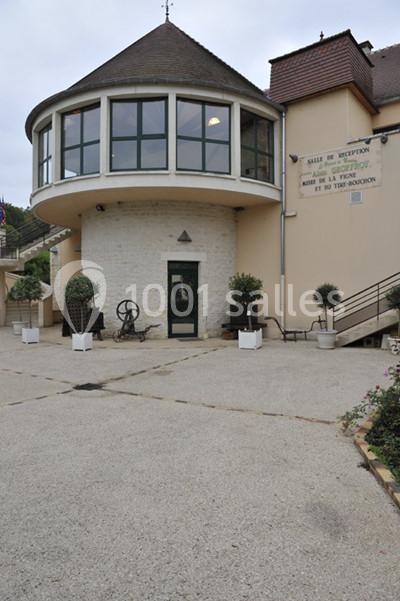Façade d'un bâtiment avec une entrée vitrée, une rampe d'accès et des jardinières sur une cour pavée.