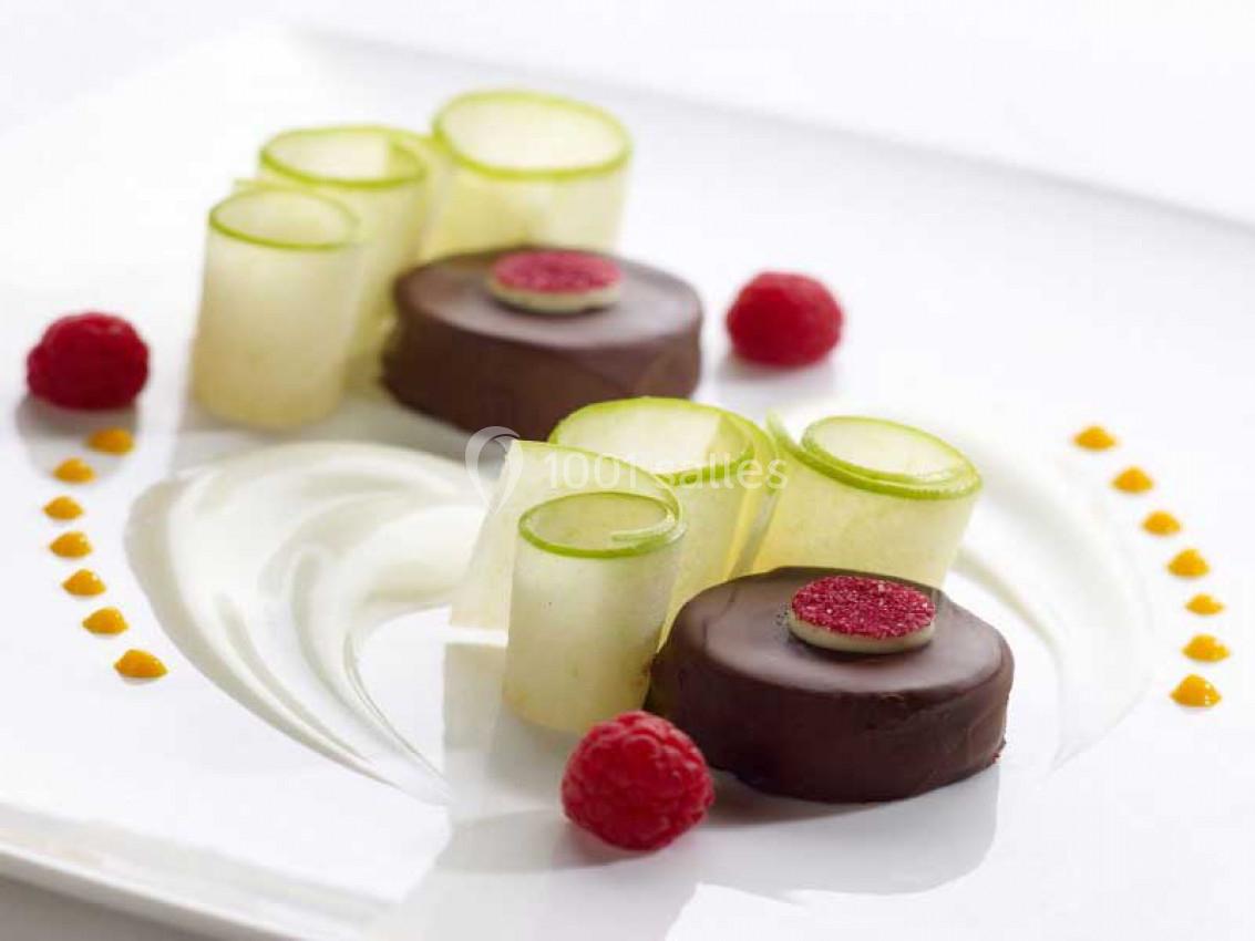 Dessert raffiné avec rondelles de pomme verte, framboises et disques en chocolat sur une assiette blanche.