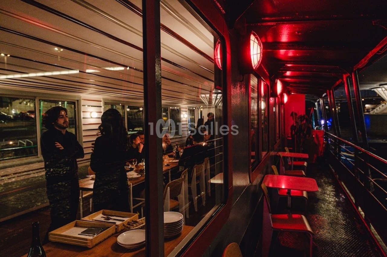 Vue d'une terrasse couverte et éclairée en rouge, avec des convives à l'intérieur d'un espace vitré adjacent.
