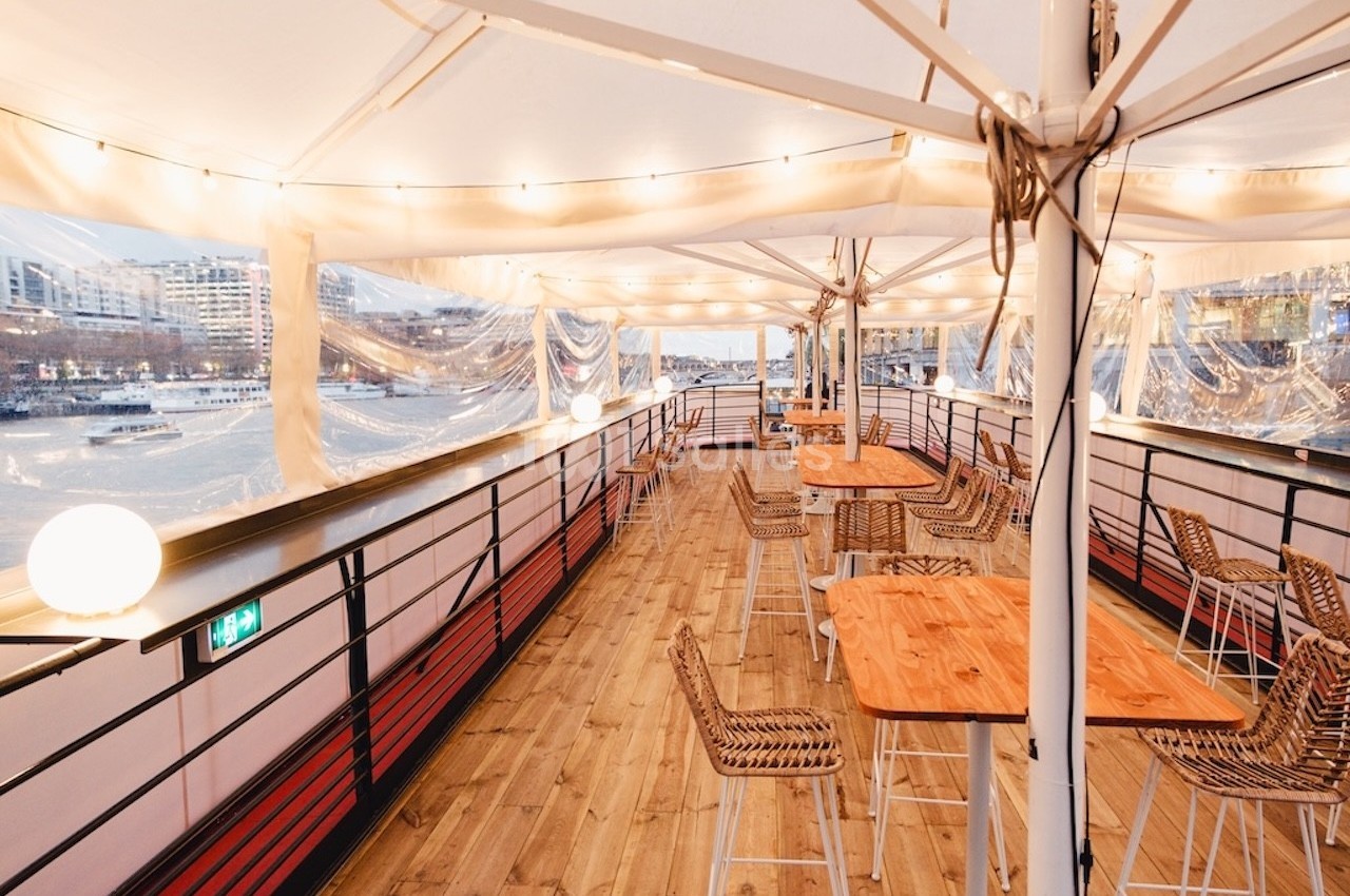Terrasse couverte d'un bateau avec tables en bois, chaises en rotin et vue sur une rivière bordée de bâtiments.