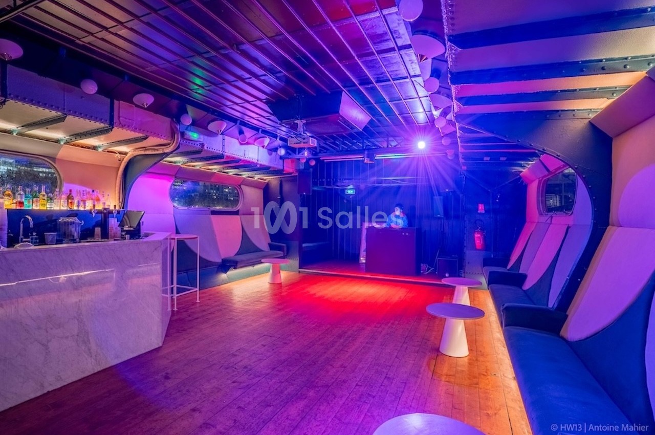 Salle de club vide avec un bar à gauche, des banquettes, un DJ au fond et un éclairage coloré.