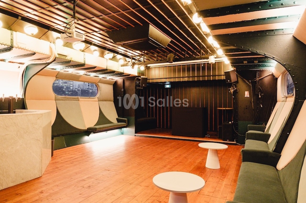 Salle intérieure moderne avec parquet, banquettes rembourrées, tables basses blanches et éclairage chaleureux.