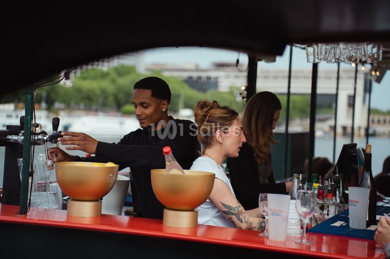 Des personnes préparent des boissons derrière un bar en extérieur, avec une rivière et des arbres en arrière-plan.