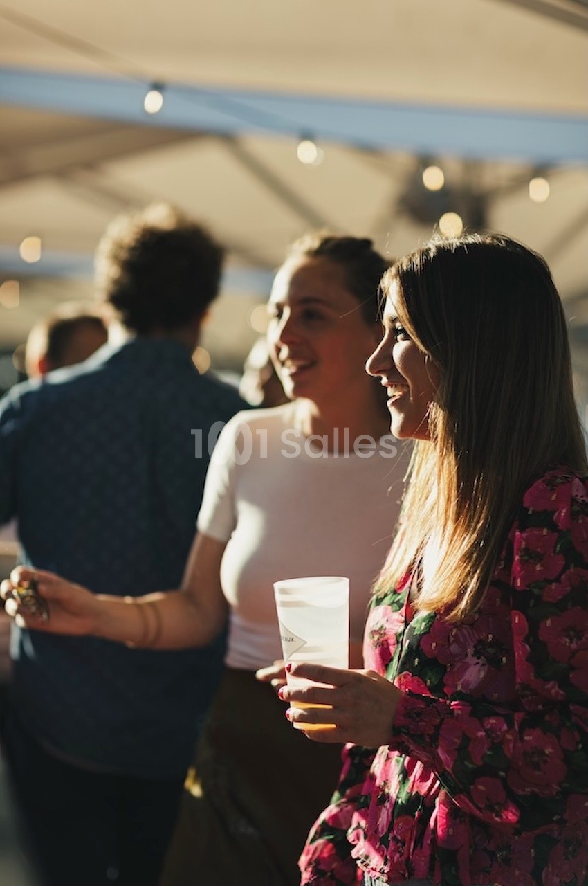 Deux femmes discutent et sourient dans un espace extérieur lumineux, tenant des boissons à la main.