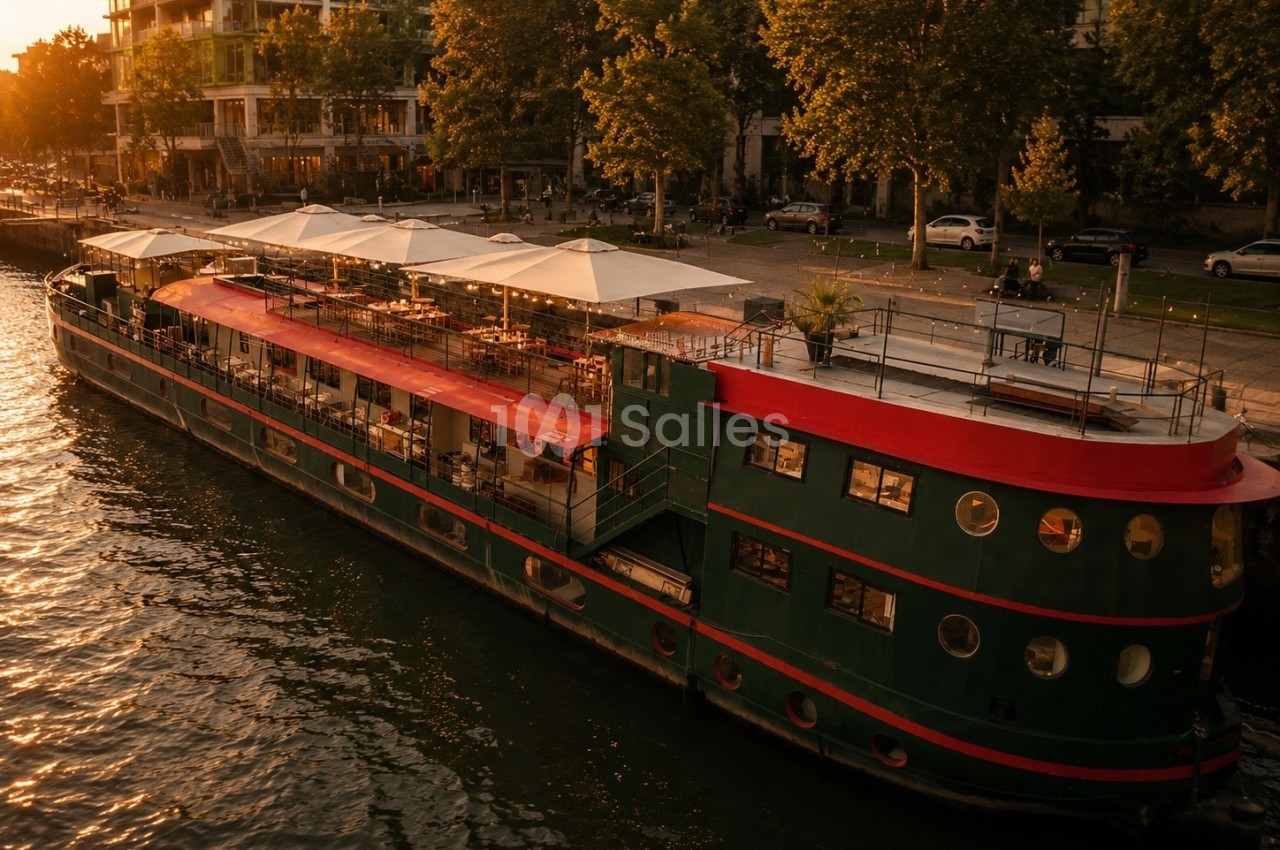 Bateau amarré au bord d'une rivière, avec une terrasse aménagée sous des parasols, entouré d'arbres et de bâtiments.