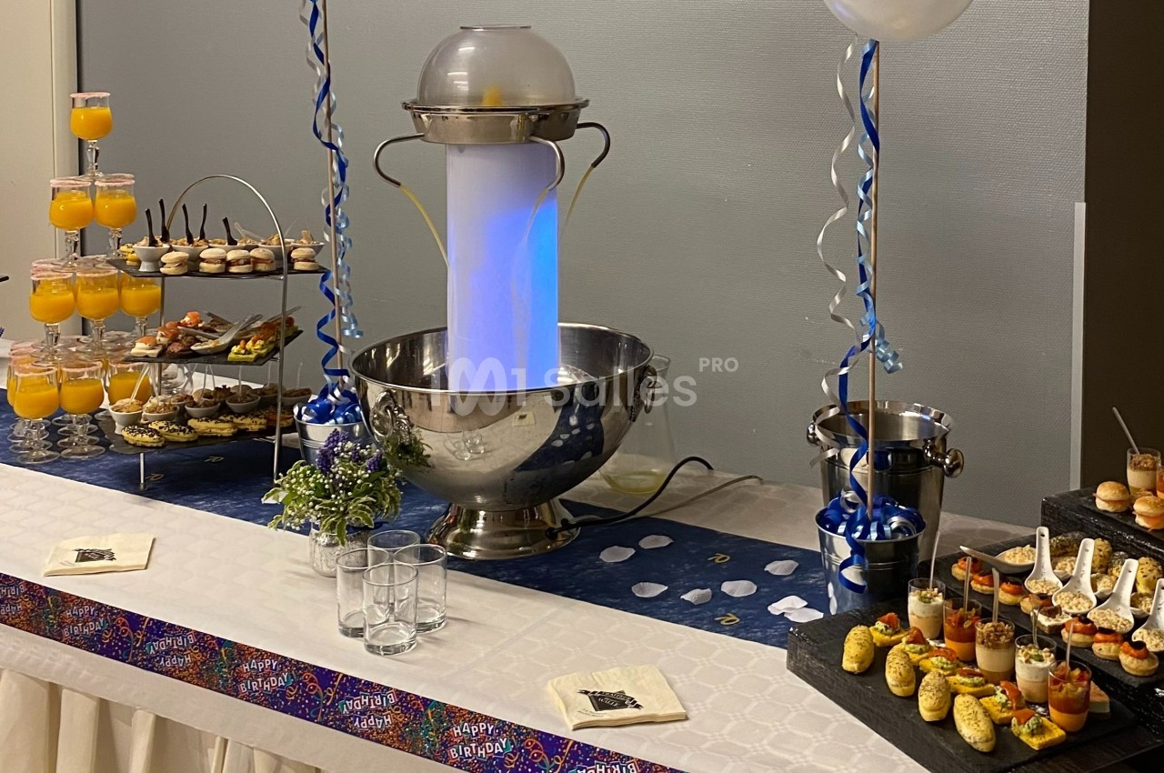 Buffet décoré avec fontaine lumineuse, verrines, amuse-bouches et boissons sur une table ornée de rubans.