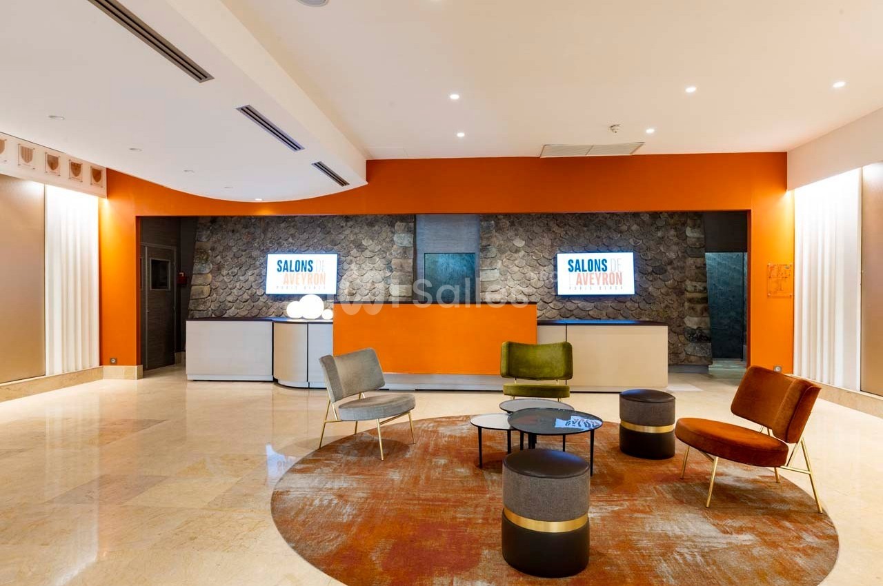 Espace d'accueil moderne avec comptoir orange, sièges colorés, tables basses et écrans affichant ’Salons Avignon’.