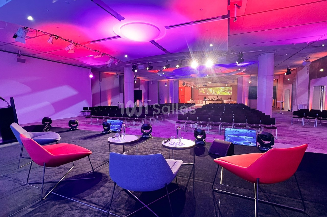 Salle de conférence moderne avec chaises colorées sur scène, éclairage violet et rangées de sièges vides en arrière-plan.