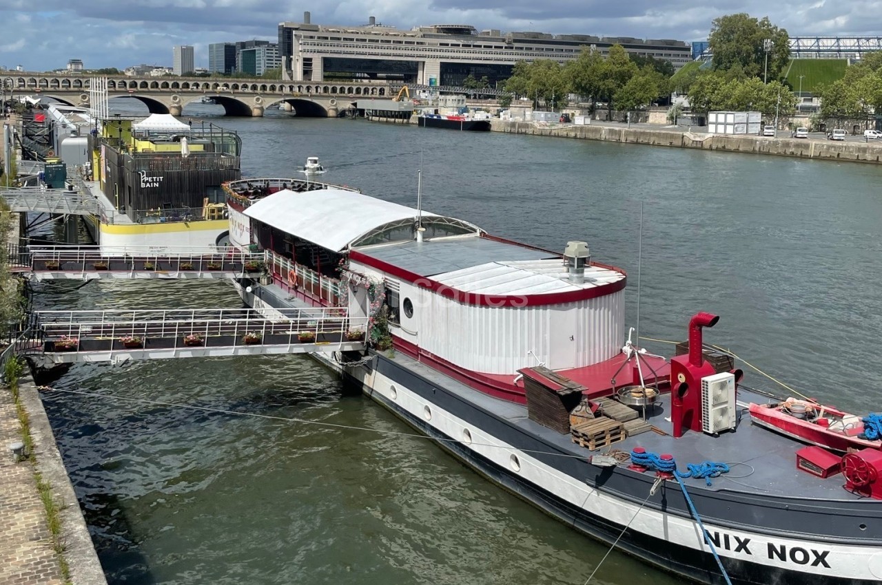Bateau blanc et rouge amarré au quai de la Seine avec chapiteau blanc, pont de pierre et bâtiments en arrière-plan.