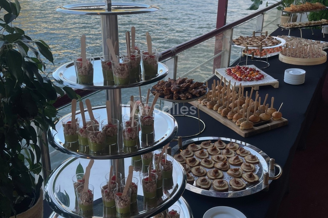 Buffet de réception en bord de fleuve avec verrines, brochettes, canapés et pâtisseries sur tables noires.
