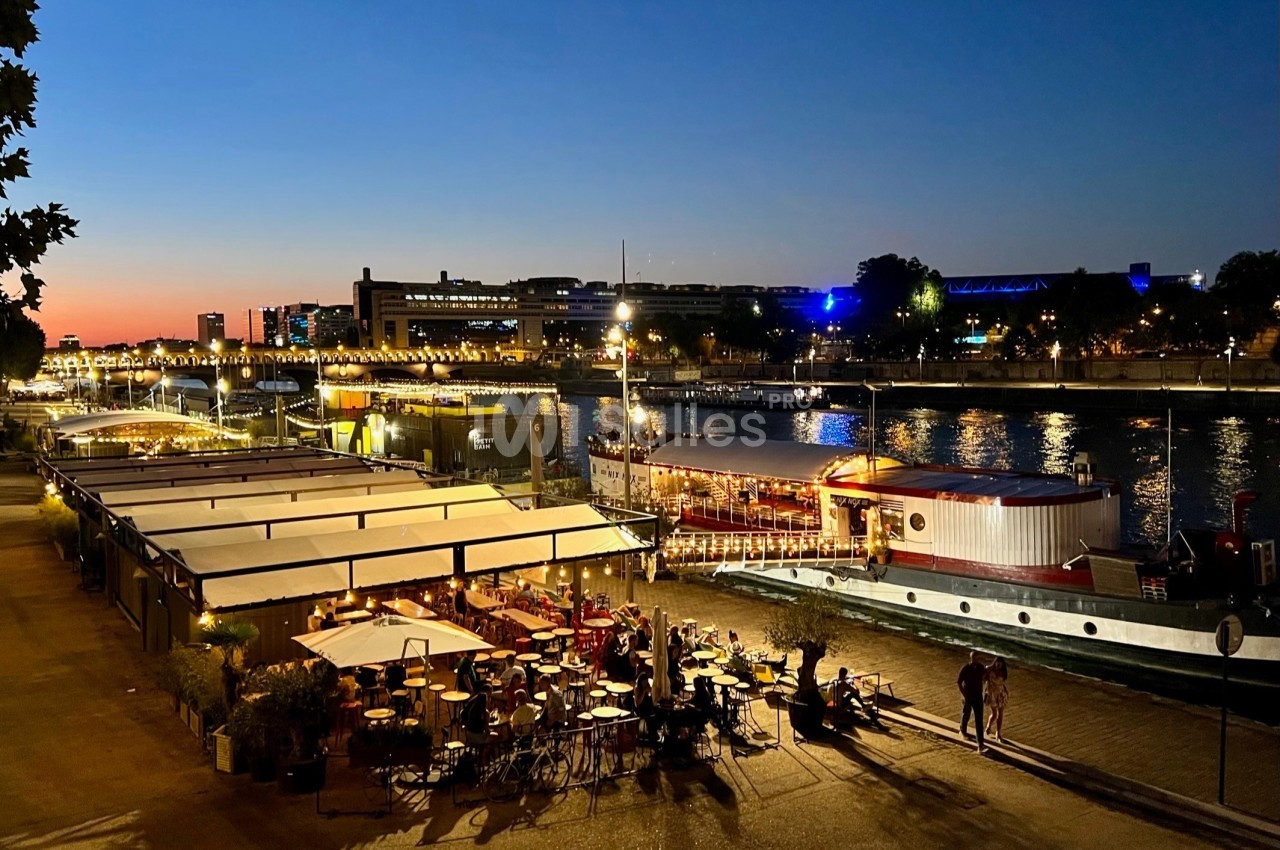 Terrasse extérieure en bord de Seine avec voile tendue, convives attablés et bateau amarré à proximité en soirée.