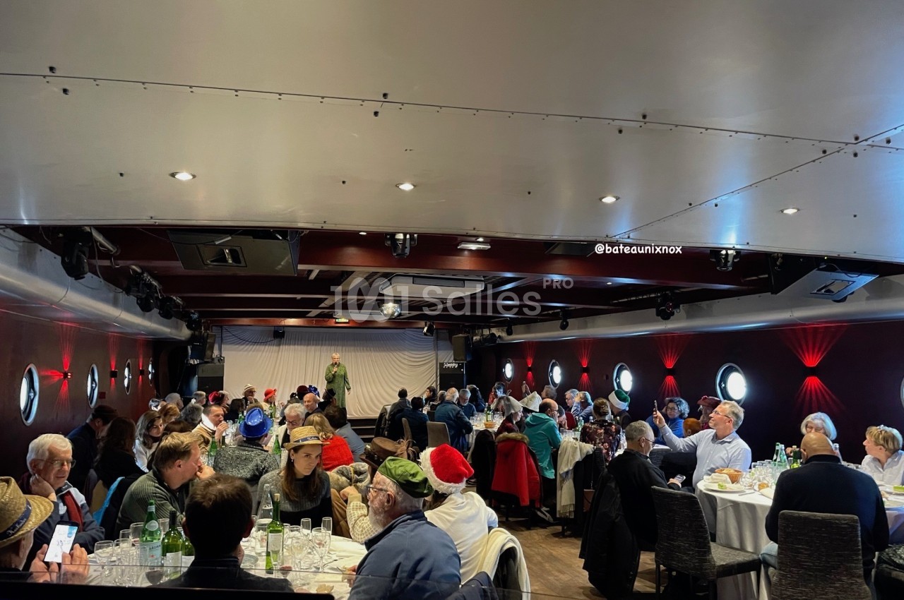 Salle intérieure dressée avec tables rondes, convives attablés et scène éclairée de projecteurs rouges.