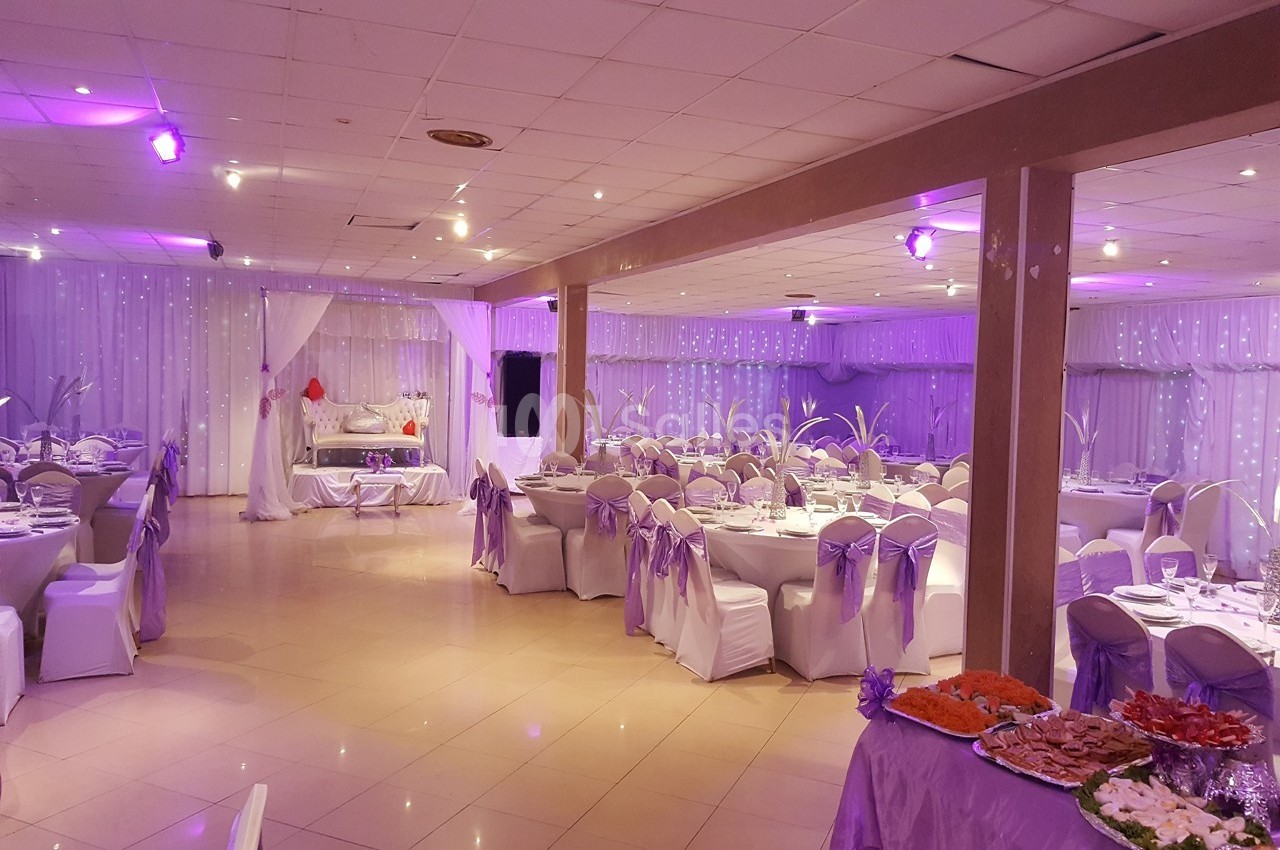Salle de réception décorée avec des nappes blanches, des rubans violets et des tables dressées pour un événement.