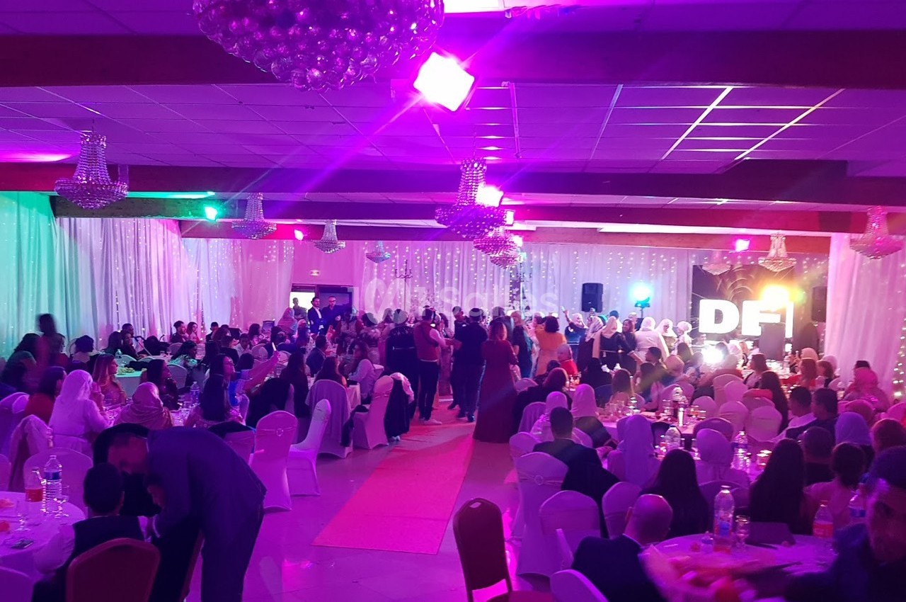 Salle de réception décorée avec des lumières colorées, remplie d'invités assis et debout lors d'un événement festif.