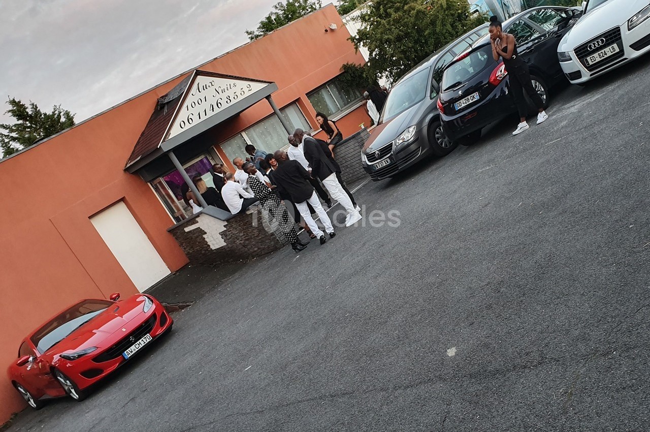 Des personnes discutent devant un bâtiment avec des voitures garées, dont une sportive rouge au premier plan.