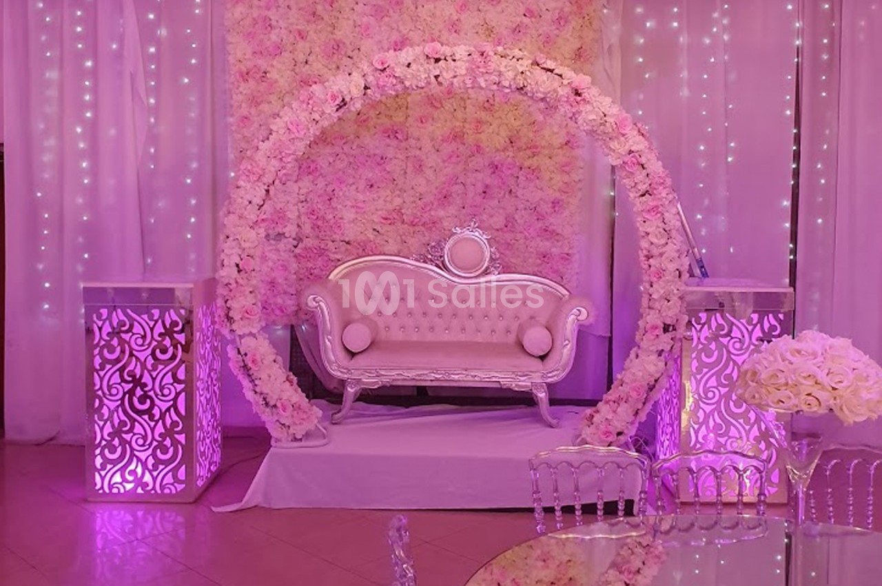 Canapé décoratif devant un mur de fleurs roses, entouré d'un arc floral et éclairé par des lumières violettes.