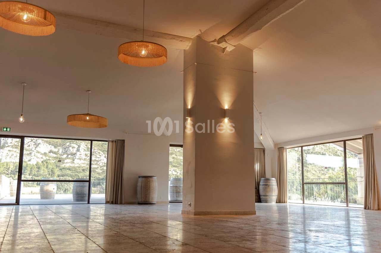 Salle lumineuse avec de grandes baies vitrées, murs clairs, poutres apparentes et suspensions en osier.