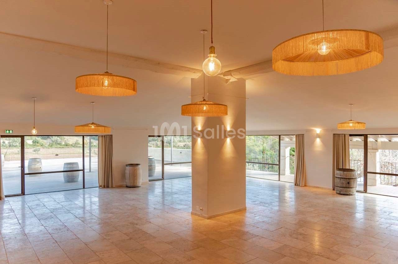 Salle lumineuse avec grandes baies vitrées, sol en pierre claire, suspensions en rotin et tonneaux décoratifs.