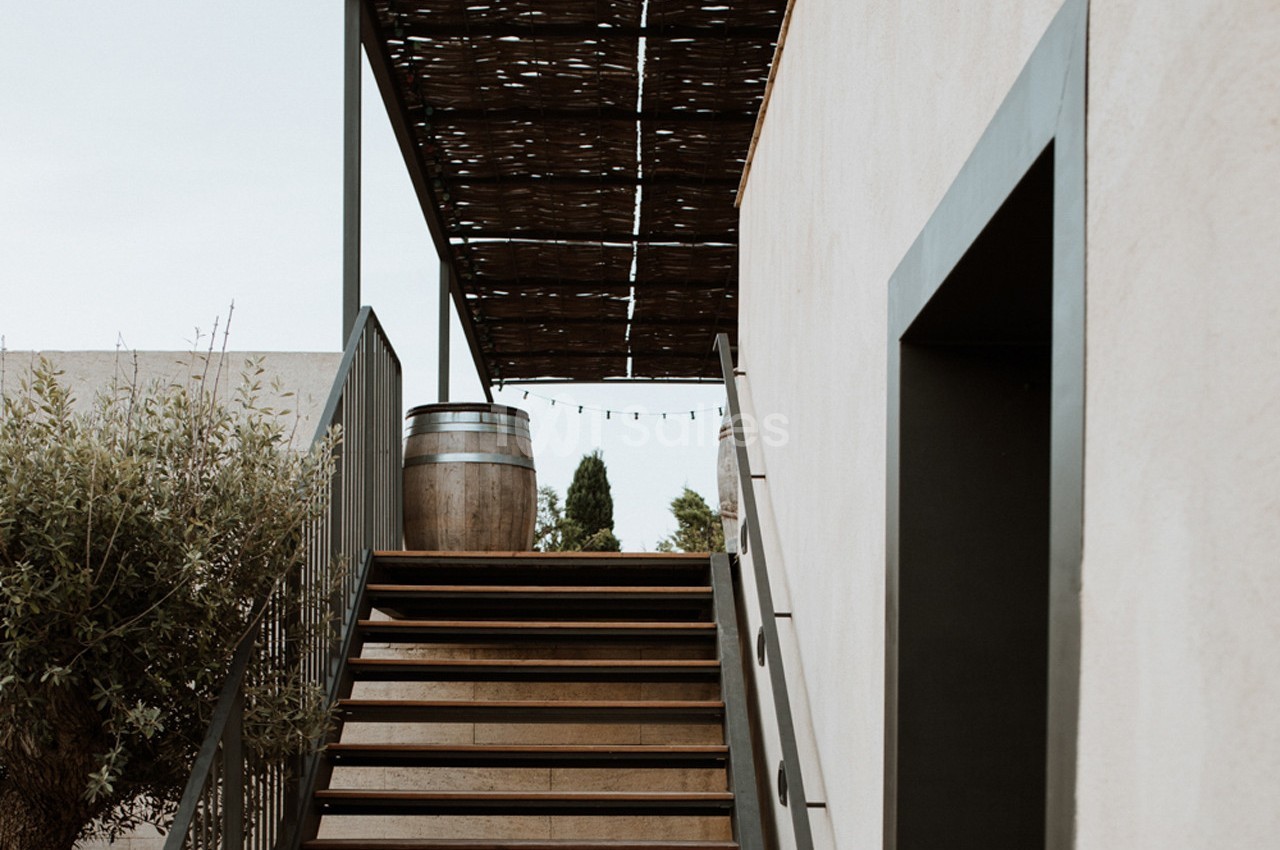 Escalier extérieur en bois menant à une terrasse couverte, avec un tonneau et des guirlandes lumineuses.