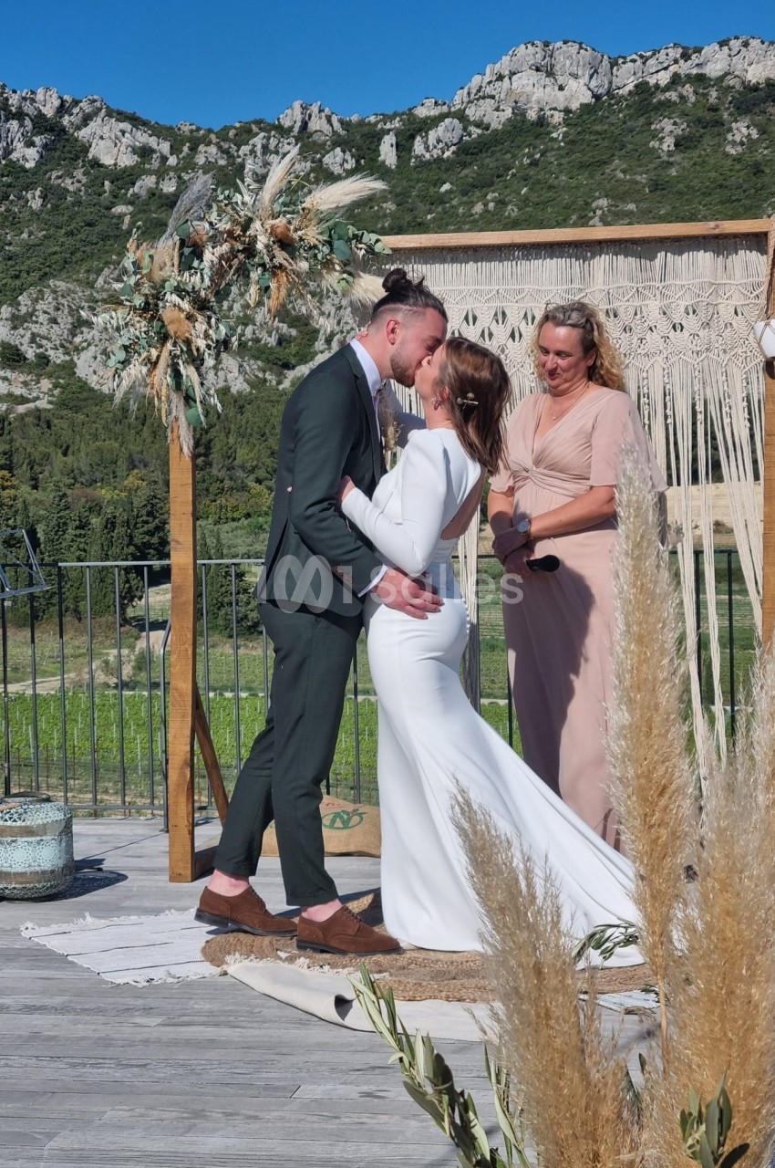Un couple s'embrasse lors d'une cérémonie de mariage en extérieur, devant un décor naturel et boisé.