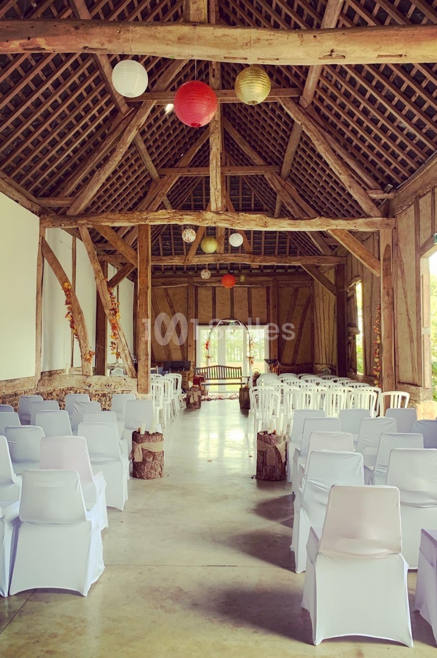Salle de réception rustique avec poutres apparentes, chaises blanches alignées et décorations suspendues.