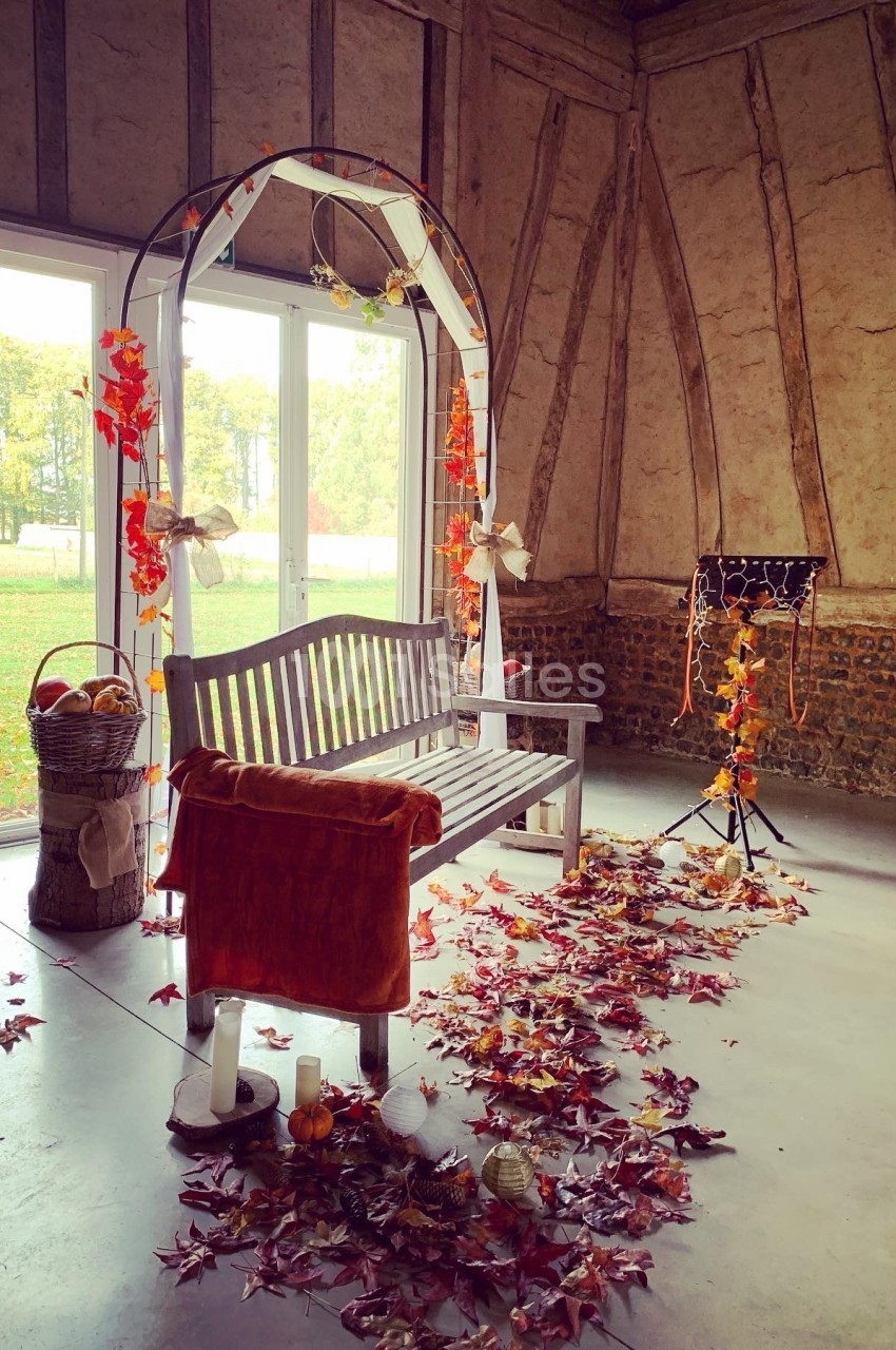 Banc en bois sous une arche décorée de feuilles d'automne, entouré de bougies et de feuilles au sol dans une salle rustique.