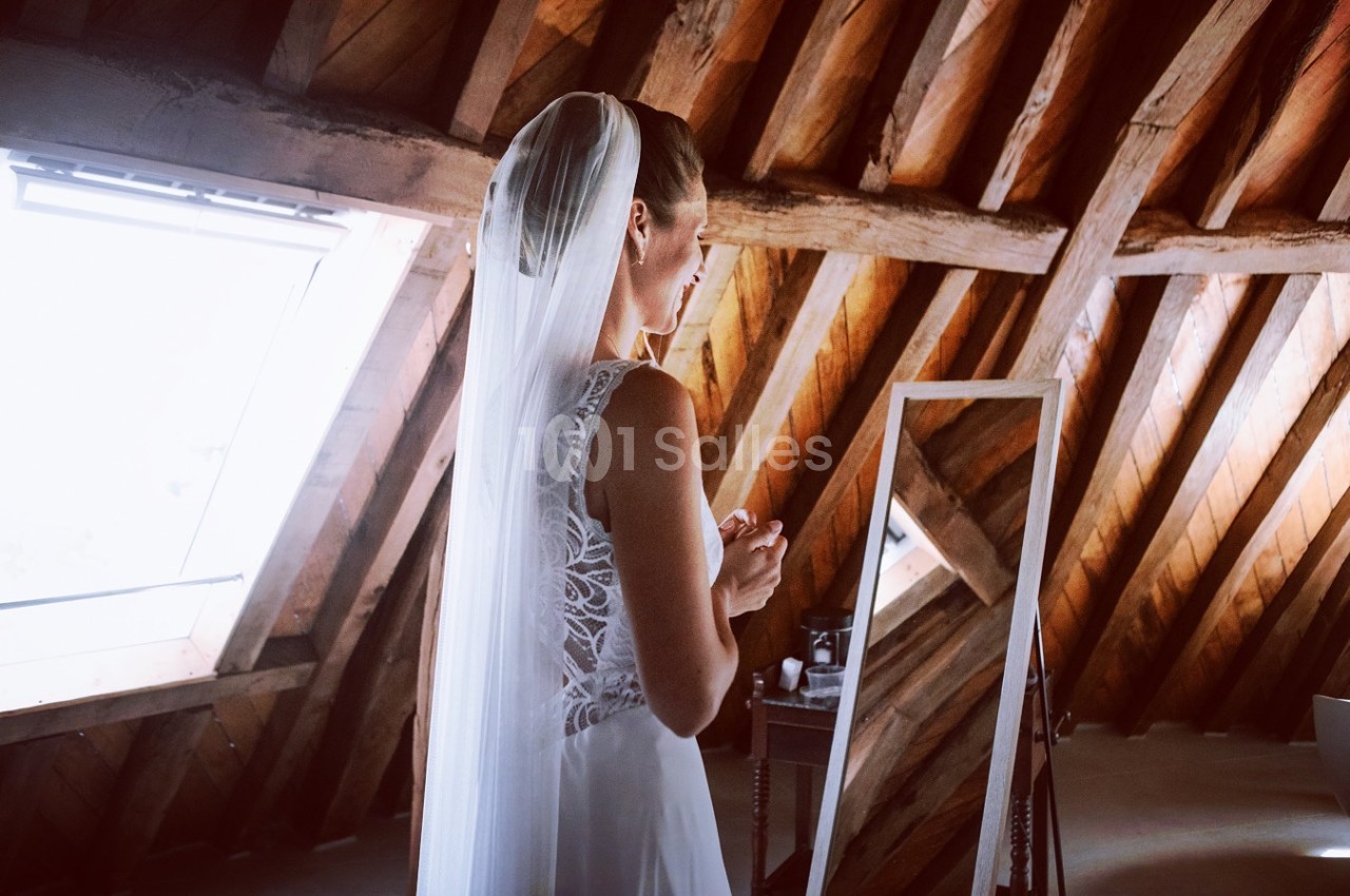 Une mariée en robe blanche et voile se prépare devant un miroir dans une pièce aux poutres apparentes.