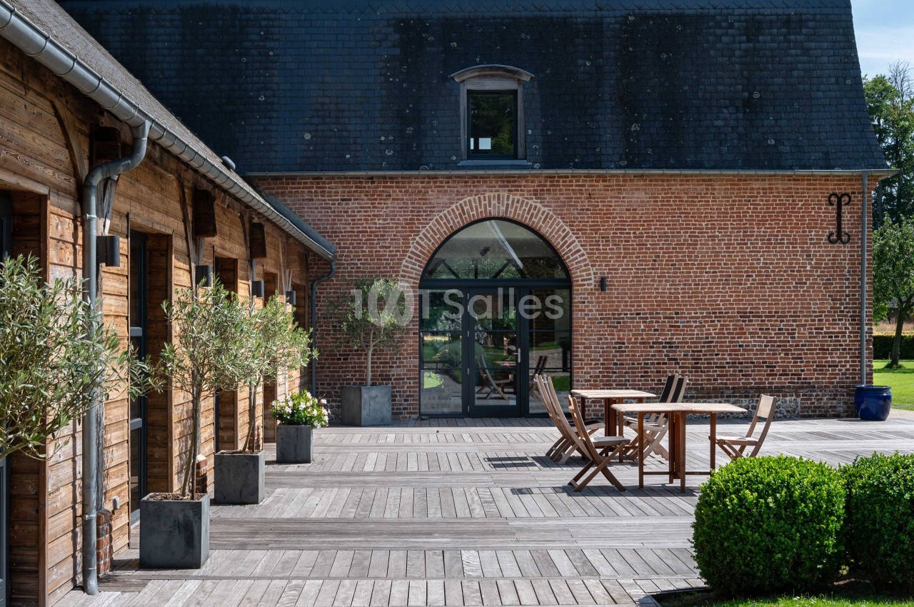 Cour en bois avec terrasse aménagée, chaises et table, entourée de bâtiments en briques et bois sous un ciel dégagé.