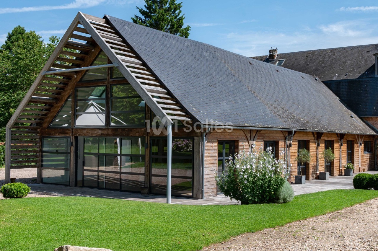 Bâtiment moderne en bois et verre avec toit en ardoise, entouré de pelouse et de végétation.