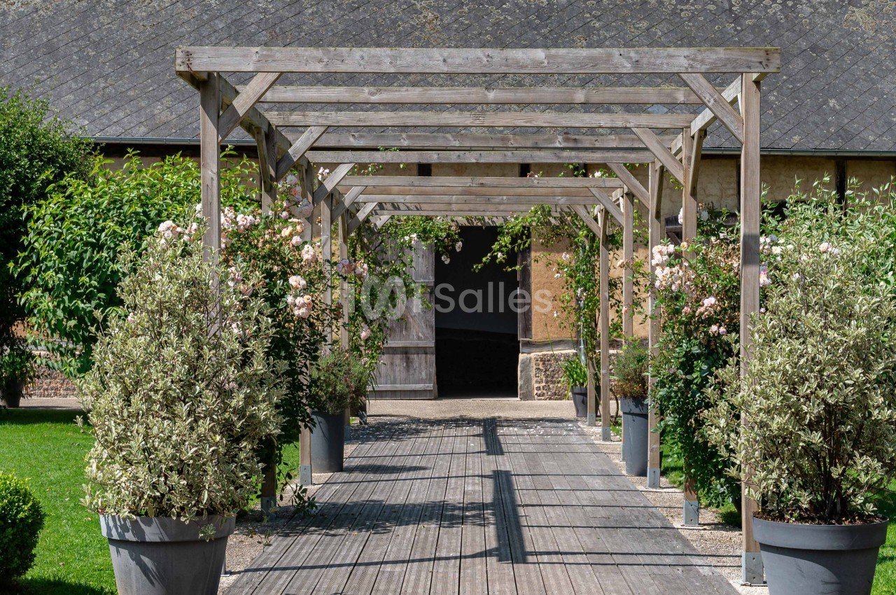 Allée en bois bordée de pots de plantes et de rosiers, menant à un bâtiment en pierre avec un toit en ardoise.