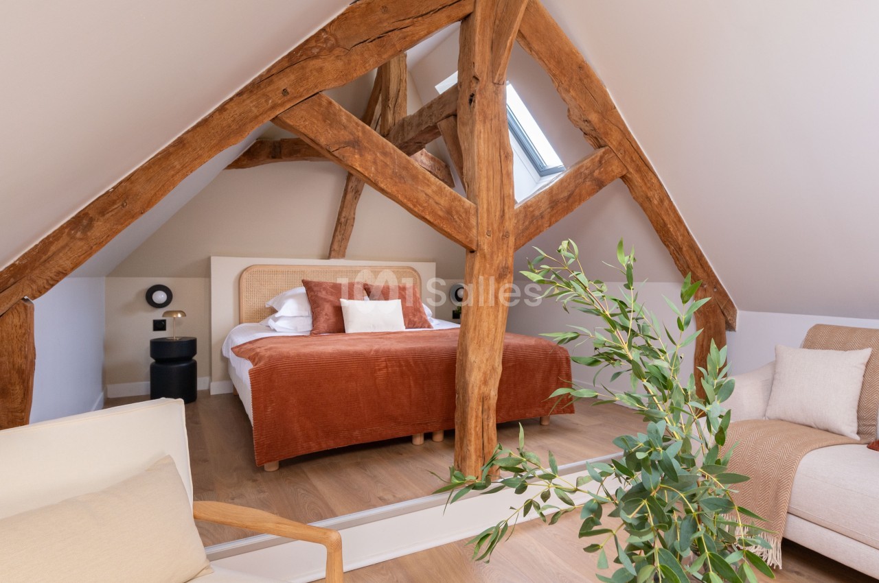 Chambre sous combles avec poutres apparentes, lit double, fauteuil beige, canapé et table basse avec plante.