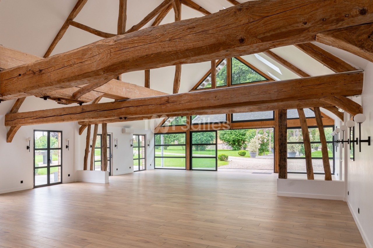 Grande pièce lumineuse avec poutres apparentes, parquet en bois clair, baies vitrées et vue sur un jardin verdoyant.